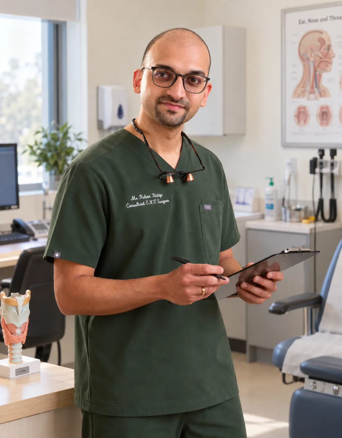 Mr Rohan Bidaye in clinic with a laryngeal model and ENT anatomy chart